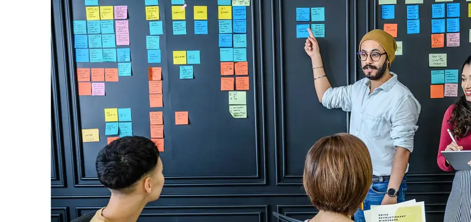 Three seated people are looking at a wall covered with post it notes, set out in columns, while a man stands and points to the notes explaining 